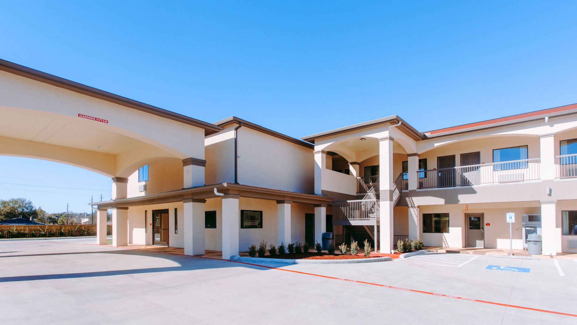 The image features a two-story building with an archway and outdoor walkways, resembling a motel or apartment complex, under a clear blue sky.