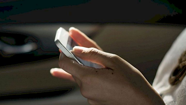 A person is holding and using a smartphone while sitting, with their hands clearly visible and the background slightly blurred.