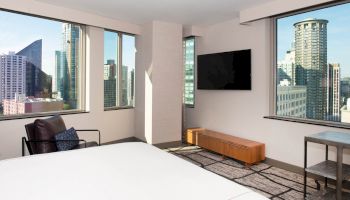 A modern hotel room with large windows offering a cityscape view, a TV, a chair, and a bed, ending the sentence.