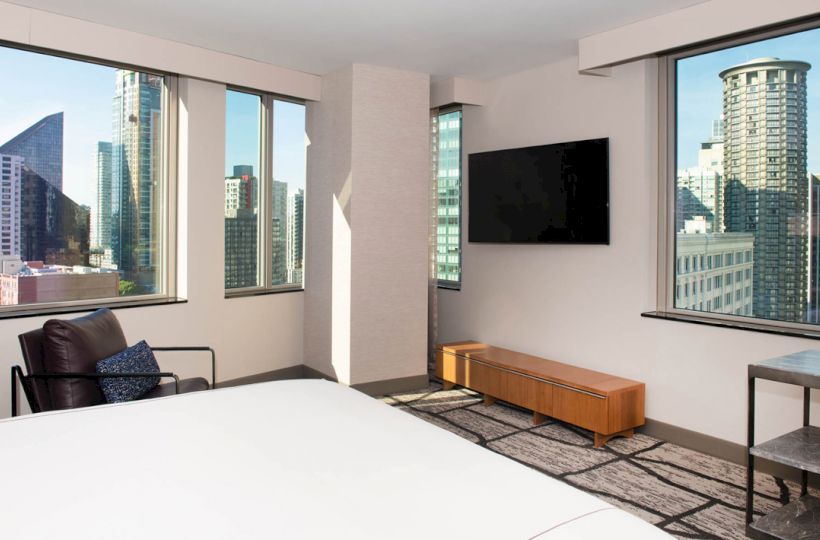 A modern hotel room with large windows offering a cityscape view, a TV, a chair, and a bed, ending the sentence.