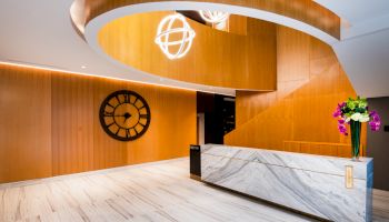 A modern interior with a marble reception desk, a large wall clock, and contemporary lighting fixtures. There are flowers on the desk.