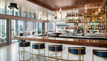 The image shows a modern bar with a wood-topped counter, blue barstools, and shelves of assorted alcohol, amid an airy space with large windows.