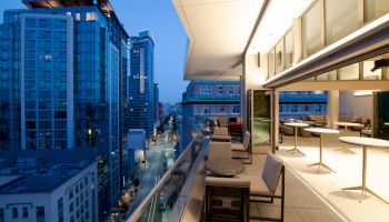 This image shows a modern urban balcony overlooking a cityscape with tall buildings at dusk. It has seating arrangements and open-air dining.