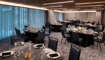 A banquet hall with round tables is set up with plates, utensils, glasses, black napkins, and floral centerpieces under modern lighting.