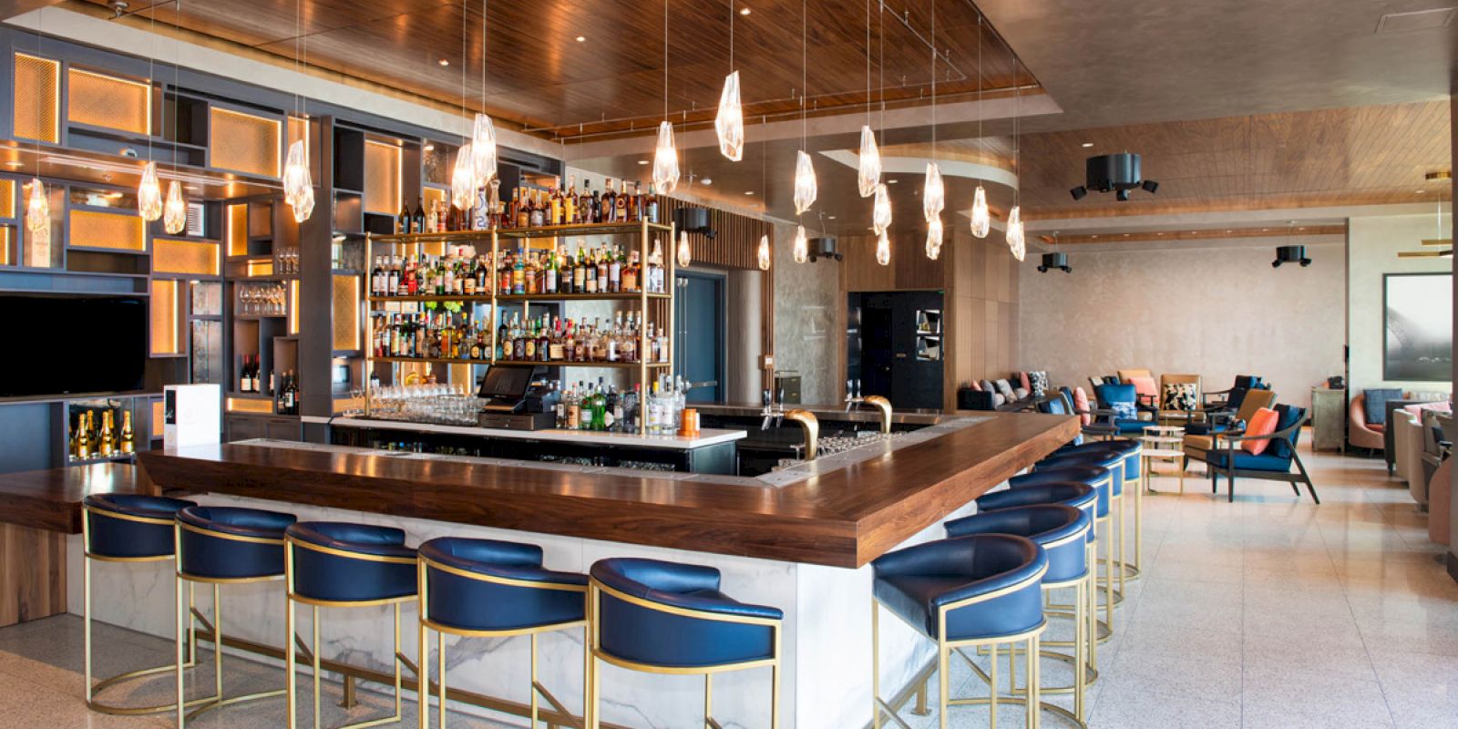 The image shows a modern bar with a wooden countertop, blue cushioned bar stools, hanging pendant lights, and a fully stocked liquor shelf.