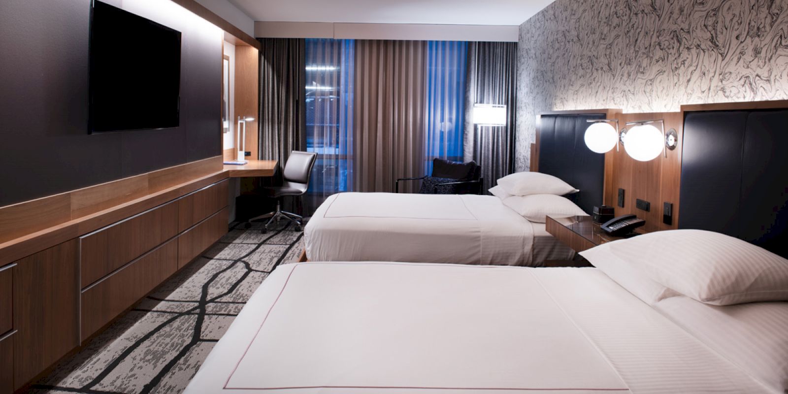 A modern hotel room features two neatly made beds, a wall-mounted TV, a desk with a chair, and stylish lighting.