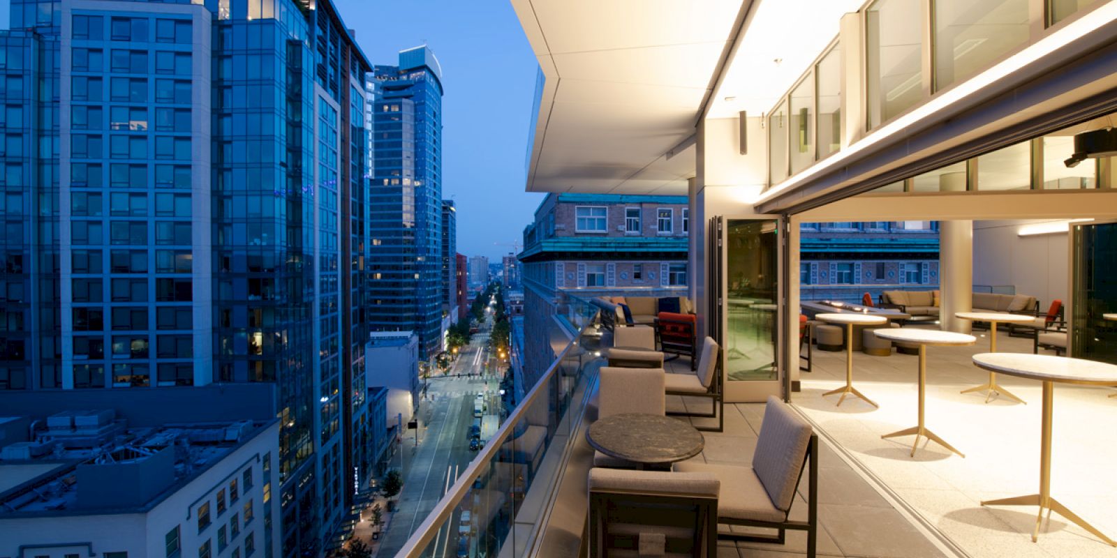 An outdoor balcony at dusk, furnished with tables and chairs, overlooks a cityscape with tall buildings and streetlights illuminating the scene.