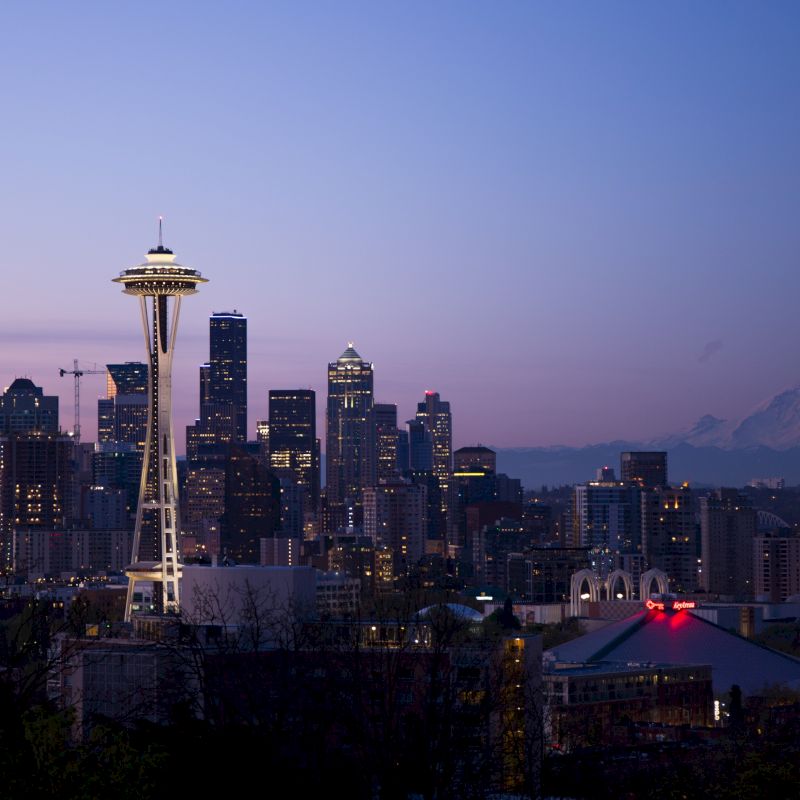 The image shows the Seattle skyline at dusk, featuring the Space Needle and Mount Rainier in the background, with city lights beginning to illuminate.