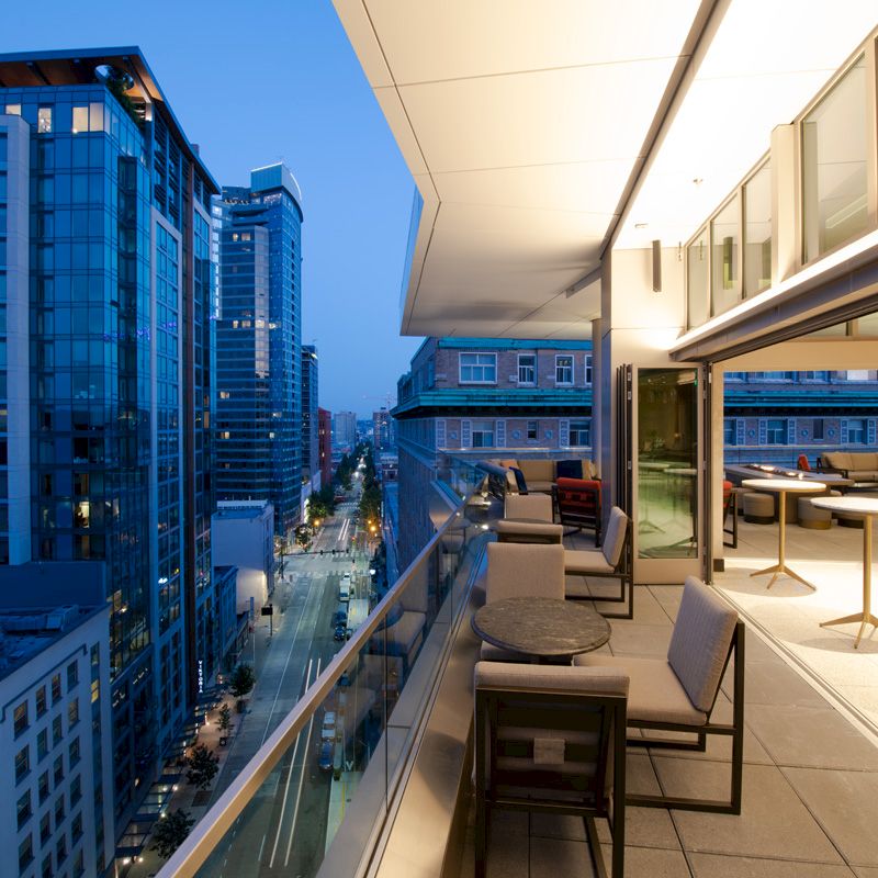 A modern cityscape view from a high-rise balcony with sleek outdoor furniture and open sliding doors leading to an indoor area.
