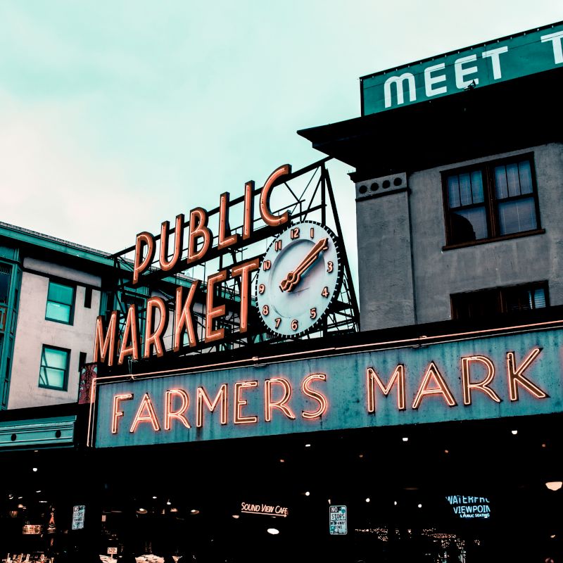 The image shows an iconic public market with neon signs for "Farmers Market" and a clock, captured against a backdrop of buildings and a cloudy sky.
