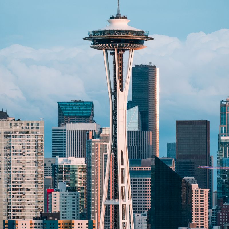 The image shows a skyline with the Space Needle centered, surrounded by various skyscrapers under a partly cloudy sky.
