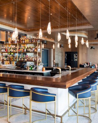 A modern bar with a variety of bottles on display, blue bar stools with gold legs, overhead pendant lights, and a cozy seating area in the background.