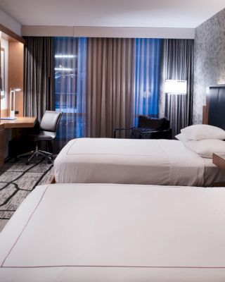 A modern hotel room features two neatly made beds, a wall-mounted TV, a wooden desk with a chair, and floor-to-ceiling windows with curtains.