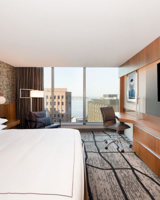 A modern hotel room features a large bed, wall-mounted TV, desk, and chair. There is a large window with a city and water view in the background.