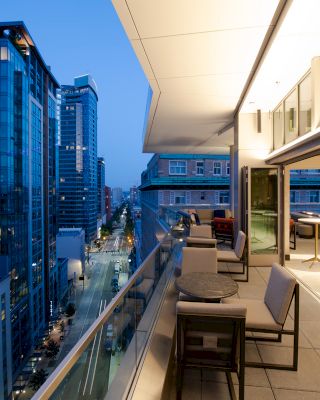 A modern urban balcony at dusk, overlooking a city street and high-rise buildings, with seating and tables on the balcony.