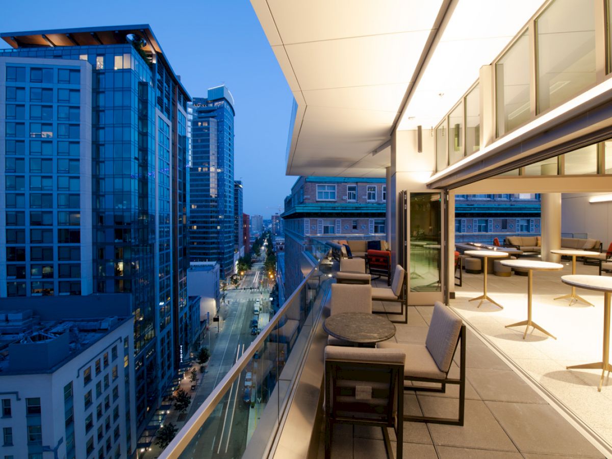 A modern cityscape at dusk from a high-rise balcony with chic outdoor seating, glass railings, and illuminated indoor spaces seamlessly connected.