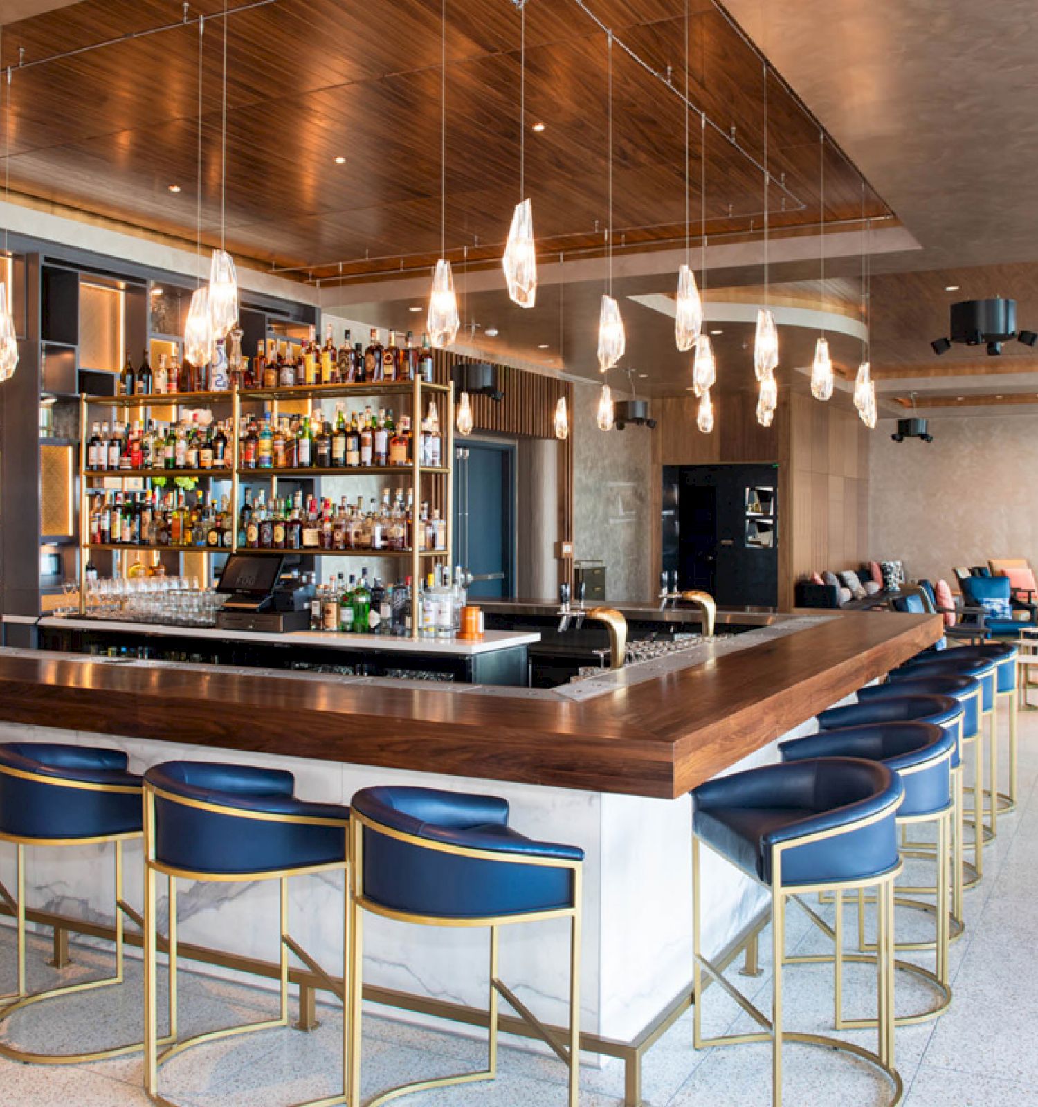 This image depicts a modern bar with blue and gold stools, a fully stocked counter, hanging lights, and a spacious seating area in the background.