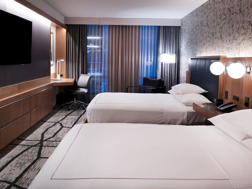 A modern hotel room with two neatly made beds, a wall-mounted TV, desk, chair, and stylish decor; large windows with curtains in the background.