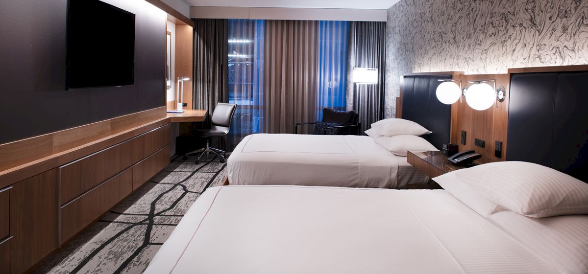 A modern hotel room with two neatly made beds, a wall-mounted TV, desk, chair, and stylish decor; large windows with curtains in the background.