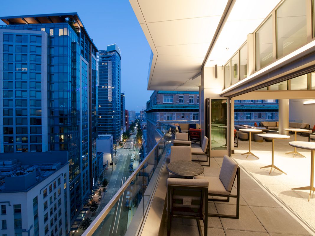 The image shows a modern cityscape at dusk from a high-rise building's balcony, featuring outdoor seating, tables, and nearby tall buildings.
