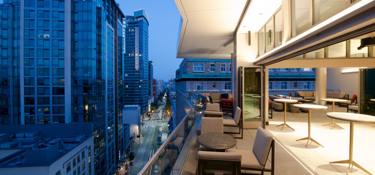 The image shows a modern cityscape at dusk from a high-rise building's balcony, featuring outdoor seating, tables, and nearby tall buildings.