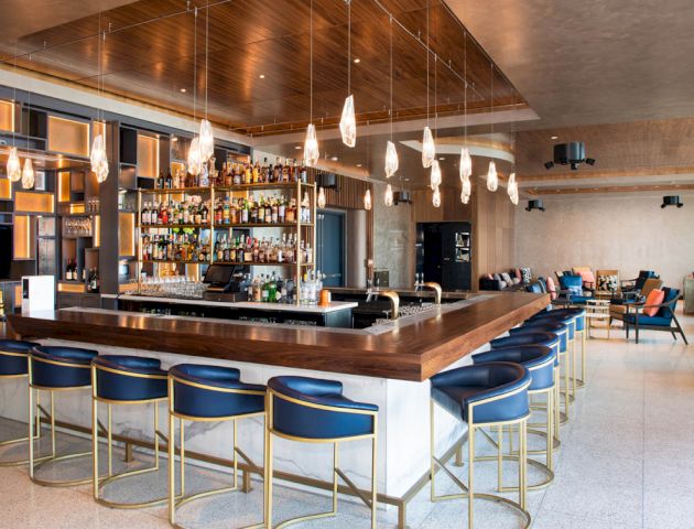 A modern bar with a range of liquor bottles, a central counter with blue and gold stools, decorative lighting, and seating areas in the background.