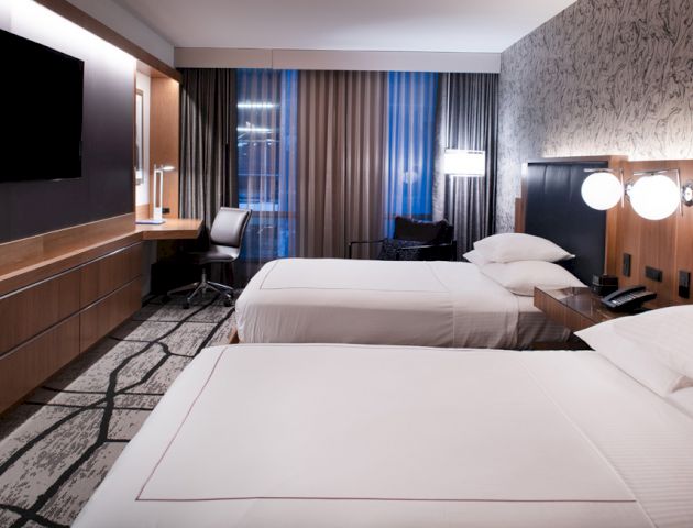 A modern hotel room with two neatly made beds, a flat-screen TV, a desk and chair, bedside lamps, and contemporary decor, including patterned wallpaper.