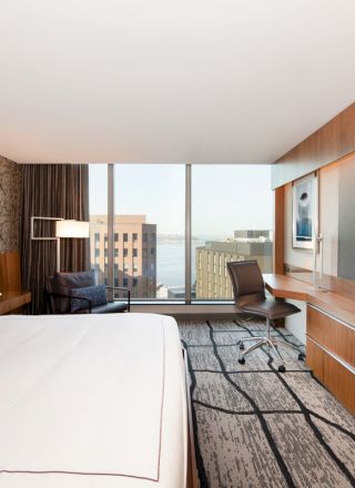 The image shows a modern hotel room with a large bed, television, desk, chair, and a view of buildings and water through a large window.