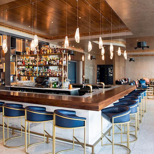 A modern, stylish bar with a wooden counter, blue barstools, hanging lights, and a fully-stocked shelf of drinks. Cozy seating area in the background.