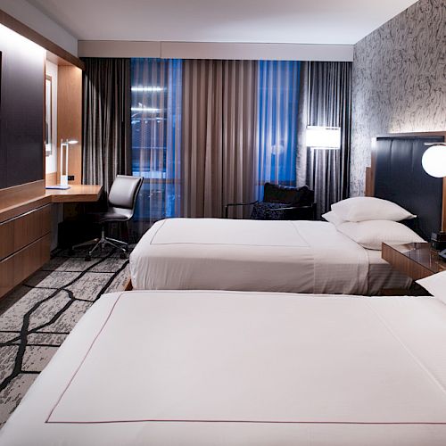 A modern hotel room with two double beds, a flat-screen TV, a desk, and a window with curtains. The room has stylish furnishings and lighting.