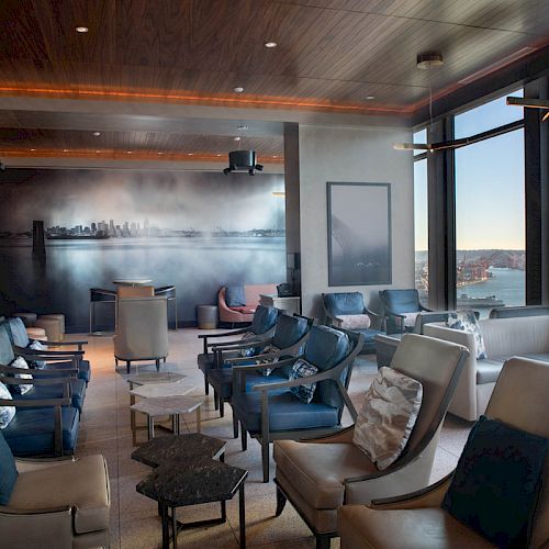 This image shows a modern lounge with numerous chairs, a scenic wall mural, and large windows providing a view of a waterfront and Ferris wheel.
