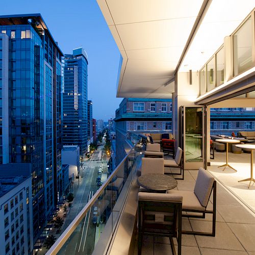 A modern outdoor patio on a high-rise building overlooks a city street and other tall buildings at dusk, featuring tables and chairs.