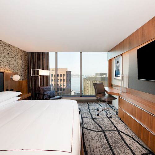This image shows a modern hotel room with a large bed, a desk and chair, a flat-screen TV, and a view of city buildings through large windows.