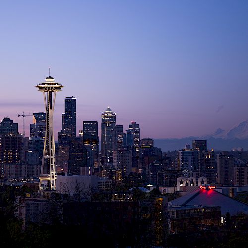 A cityscape of Seattle with the Space Needle prominently in view, tall buildings, and Mount Rainier in the background during twilight.
