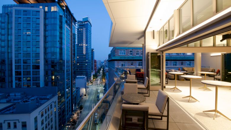 A modern cityscape at dusk viewed from an upscale balcony, featuring lounge chairs, tables, and large windows overlooking tall buildings and a lit street.