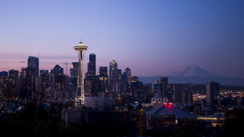 The image shows the Seattle skyline at dusk, with the Space Needle prominently visible and Mount Rainier in the background against a twilight sky.