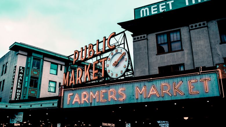 The image shows the entrance of a public farmers market with a clock. Signs for a nearby hotel and a partially visible "MEET THE" sign appear too.