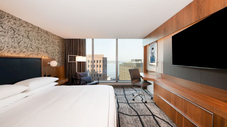 A modern hotel room with a large bed, wall-mounted TV, a desk with a chair, and a window offering a view of city buildings and a body of water.