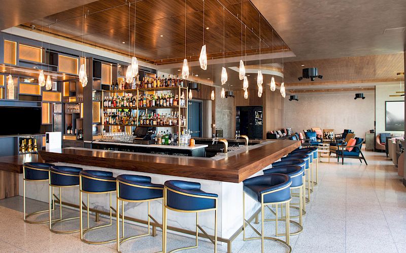 The image shows a modern bar with blue and gold stools, a central bar area stocked with various bottles, and stylish lighting fixtures hanging from the ceiling.
