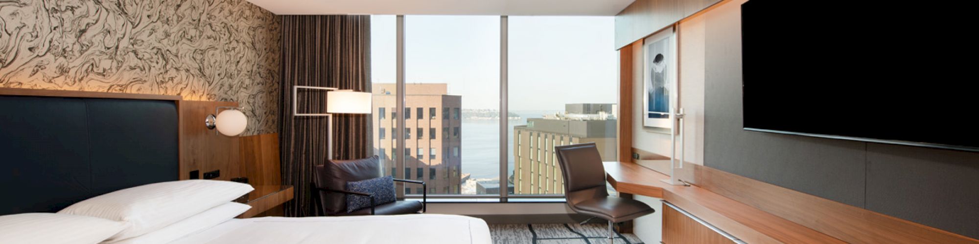 A modern hotel room features a large bed, a wall-mounted TV, a desk with a chair, large windows with a city view, and contemporary decor.